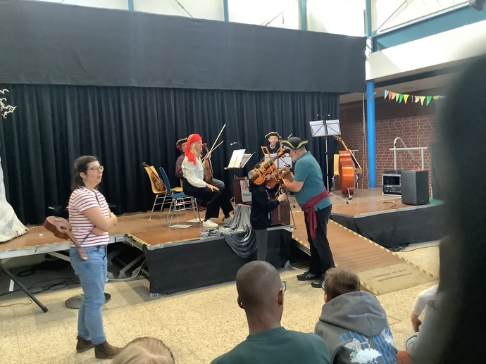 Musiker sitzen auf der Bühne. Vor der Bühne zeigt ein Musiker einer Schülerin, wie man Geige spielt.