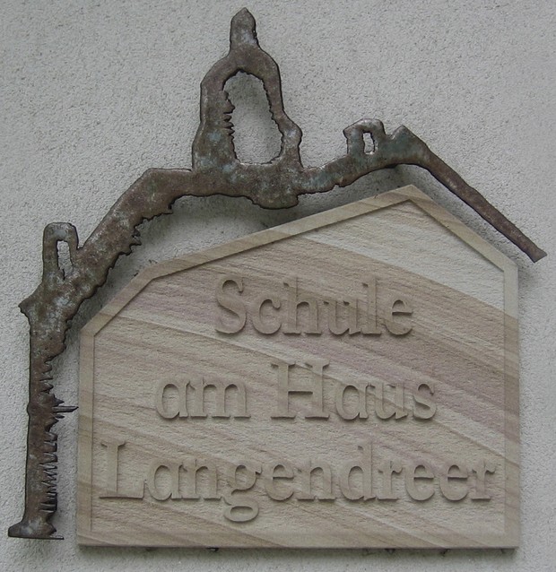 Das Foto zeigt eine Sandsteintafel mit der Aufschrift "Schule am Haus Langendreer". Über der Sandsteintafel befindet sich eine Metall-Nachbildung der Silhouette des Glockenturms vom Haus Langendreer.