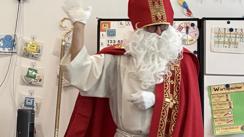 Der Nikolaus ist steht vor einer Reihe sitzender Schülerinnen und Schüler und winkt.