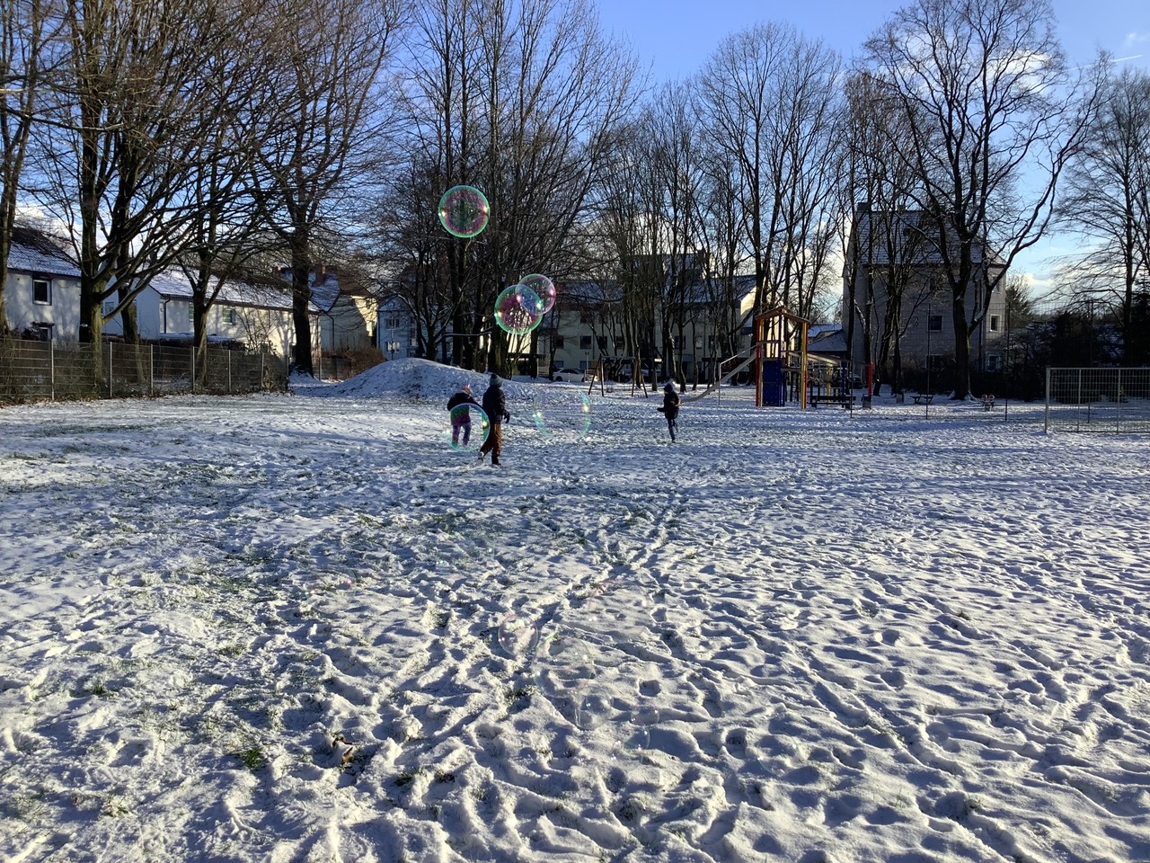 Spielplatz mit drei Kindern im Hintergrund, es liegt Schnee. im Vordergrund Seifenblasen