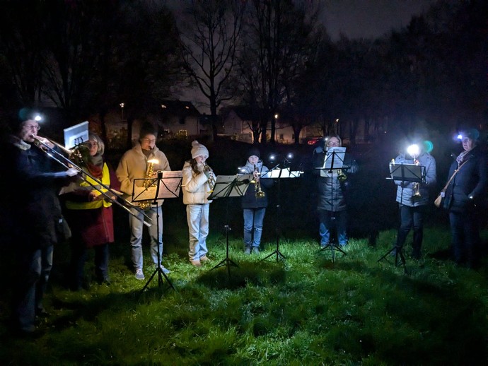 acht personen stehen im Halbkreis vor Notenständern und spielen bei Taschenlampenlicht Blasinstrumente