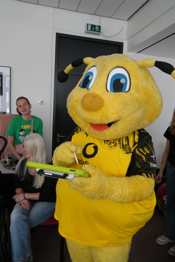 BVB Maskottchen Emma nutzt einen SPrachcomputer. Im Hintergrund scvhauen zwei Frauen zu.