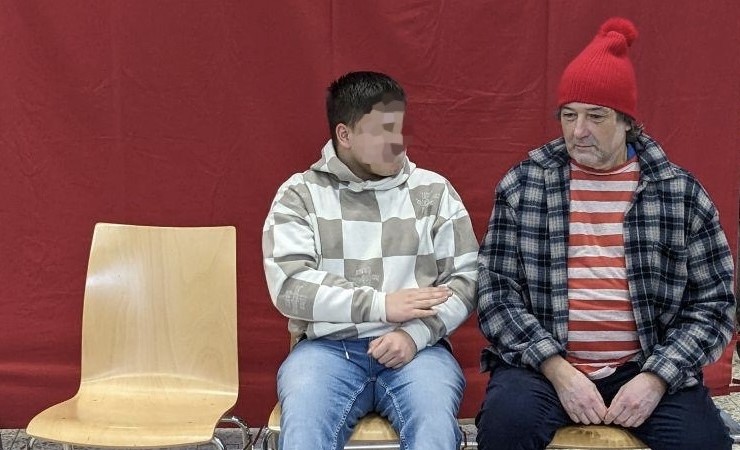 Ein Schüler und ein Mann sitzen auf Stühlen nebeneinander vor einem roten Hintergrund.