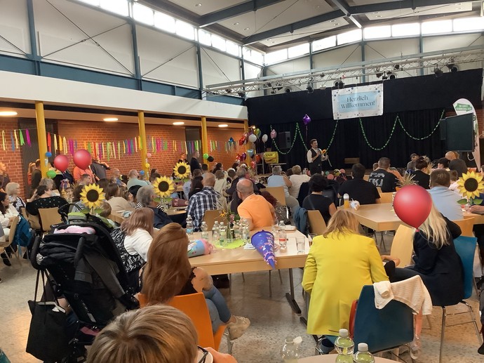 Viele Personen sitzen in einer Aula an Tischen. Ein Mann redet in ein Mikrophon.