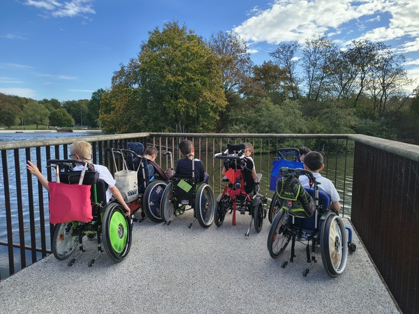 6 Kinder sitzen im Rollstuhl und schauen durch ein Geländer auf einen See, man sieht sie von hinten