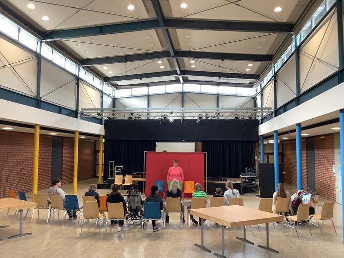 Vor einer roten Stellwand sitzen zwei Erwachsene mit Blick zum Publikum. Das Publikum ist in Rückenansicht zu sehen. (vergrößerte Bildansicht wird geöffnet)