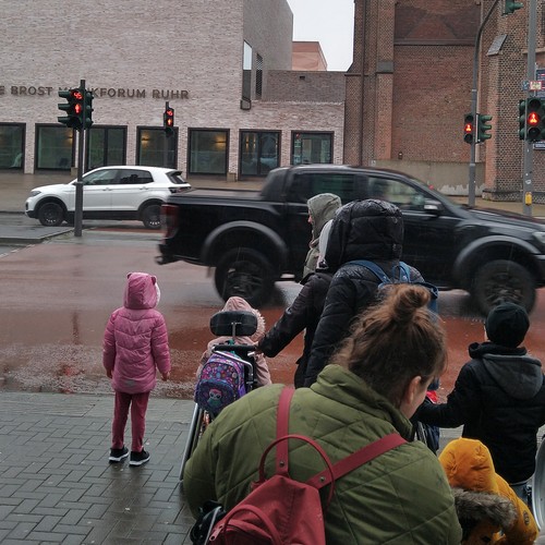 Kinder von hinten zu sehen stehen an einer Ampel. Auf der anderen Seite am Gebäude der Schriftzug "Anneliese Brost Musikforum Ruhr"
