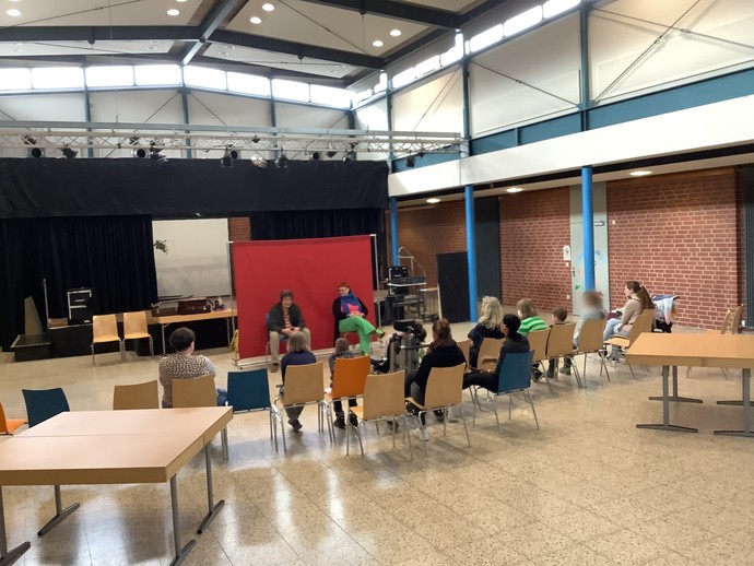 Vor einer roten Stellwand sitzen zwei Erwachsene mit Blick zum Publikum. Das Publikum ist in Rückenansicht zu sehen. (vergrößerte Bildansicht wird geöffnet)