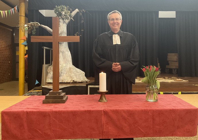 Ein evangelischer Pfarrer steht hinter einem Tisch mit roter Tischdecke. Auf dem Tisch stehen Blumen, eine Kerze und ein Kreuz.