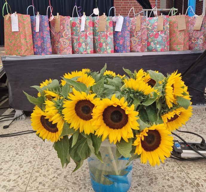 Ein Strauß Sonnenblumen steht in einer Vase, dahinter sind aufgereiht Geschenktüten zu sehen.