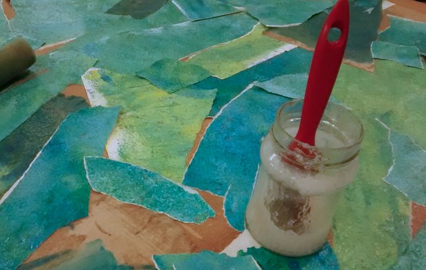 Grün und blau angemalte Papierschnipsel leigen auf dem Boden. Darauf steht ein Glas mit Kleister. Im Glas steckt ein Pinsel mit rotem Griff.