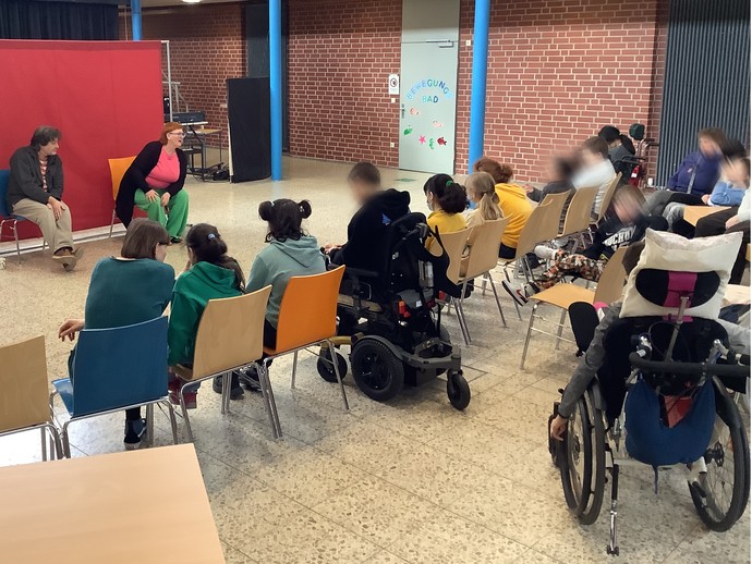 Vor einer roten Stellwand sitzen zwei Erwachsene mit Blick zum Publikum. Das Publikum ist in Rückenansicht zu sehen. (vergrößerte Bildansicht wird geöffnet)