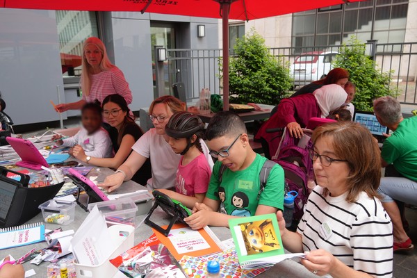 Mehrere Erwachsene und Kinder sitzen um einen Tisch, nutzen einen Sprachcomputer und basteln