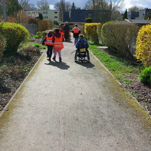 Kinder mit orangenen Warnwesten von hinten zu sehen laufen auf einem Weg durch eine Kleingartenanlage.