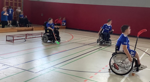 mehrere Personen in blauen Trikots in Sportrollstühlen mit Hockeschlägern in einer Turnhalle