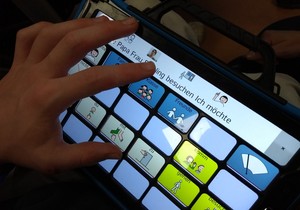 Eine Person wählt auf einem Sprachcomputer Symbole aus und stellt so einen Satz zusammen. Nur die Hand und die Bedienoberfläche sind zu sehen.