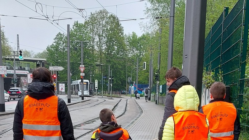 Eine Gruppe Schülerinnen und Schüler mit Warnwesten und Greifzangen stehen an Bahnschienen. Es regnet. Sie sind von hinten zu sehen.