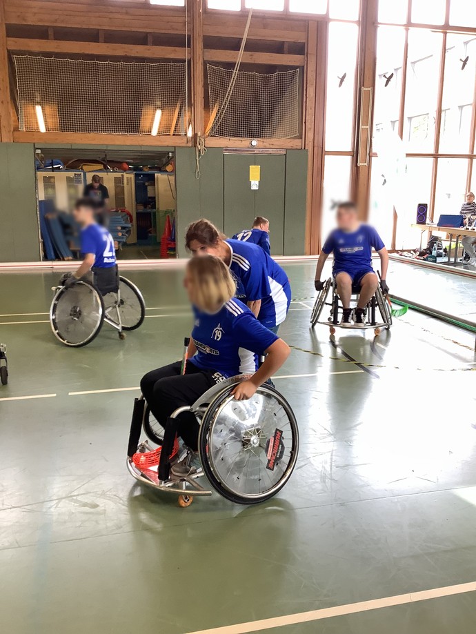Einige Aktivrollstuhl-Hockeyspielerinnen und Spieler sind auf dem Platz. Eine Frau gibt Ratschläge.