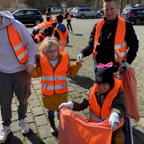 Mehrere Personen (Erwachsene und Kinder, teils im Rollstuhl sitzend) in orangenen Warnwesten mit Greifzangen und Müllbeuteln.