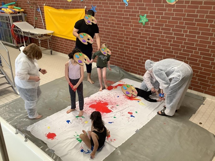 Eine Gruppe Personen in Maleranzügen malt mit dem Körper auf ein weißes Tuch.
