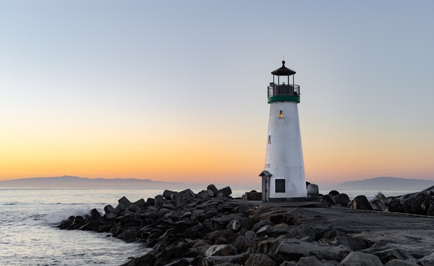 Ein weißer Leuchtturm mit schwarzem Dach steht auf einem flachen, felsigen Küstenabschnitt im Sonnenaufgang am Meer.