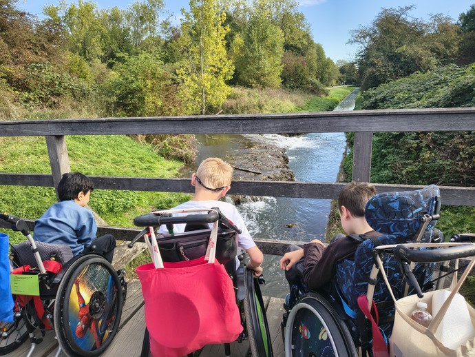 3 Kinder im Rollstuhl, man sieht sie von hinten, sie schauen durch das Brückengeländer auf einen Wasserlauf