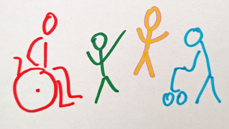 Vier Personen als gezeichnete Strichmännchen nebeneinander. Von links nach rechts: Eine rote Person im Rollstuhl, eine grüne und eine gelbe stehende Person, eine blaue mit Rollator.