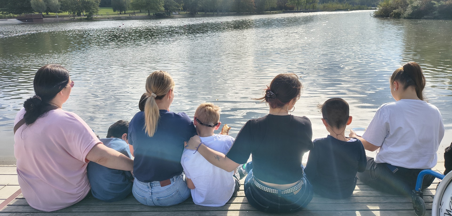 drei Kinder und vier Erwachsene sitzen auf einem Holzsteg und schauen aufs Wasser. Sie sind von hinten zu sehen.