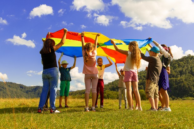Acht Kinder stehen auf einer Wiese und heben gemeinsam ein buntes Schwungtuch in die Höhe.