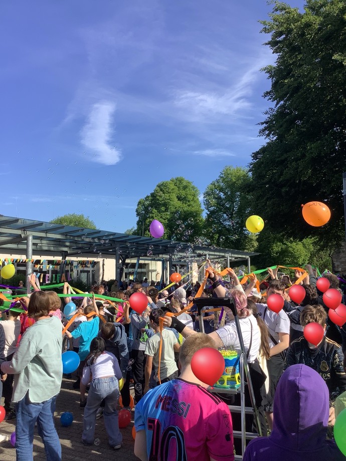 Viele Personen stehen draußen und sind verbunden mit bunten Bändern. Luftballons fliegen