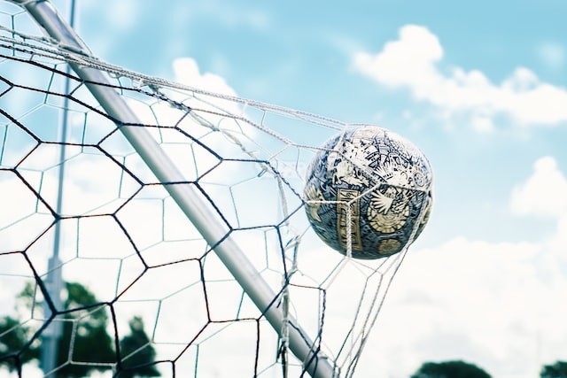 Außenansicht eines Ausschnitts eines Fußballtors: ein Lederfußball drückt vor blauem Himmel das Netz nach außen.