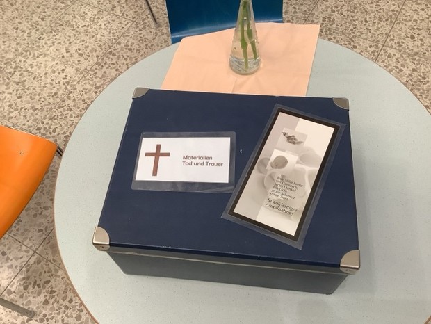 Eine blaue Pappbox mit Metallecken auf dem ein Schild mit einem Kreuz und den Worten "Materialien Tod und Trauer" klebt, daneben klebt eine Beileidskarte