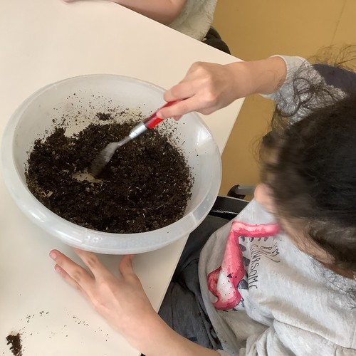 Zwei Kinder mit einer Schalle voll Erde. Ein Kind spritzt mit einer Blumenspritze auf die Erde.