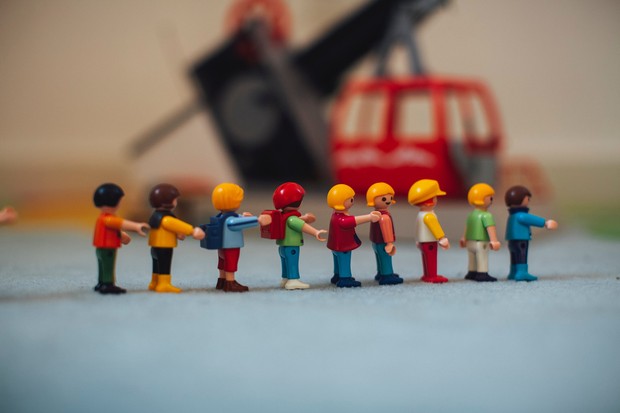 Neun bunte Playmobil-Kinderfiguren stehen mit ausgestreckten Armen hintereinander in einer Reihe.