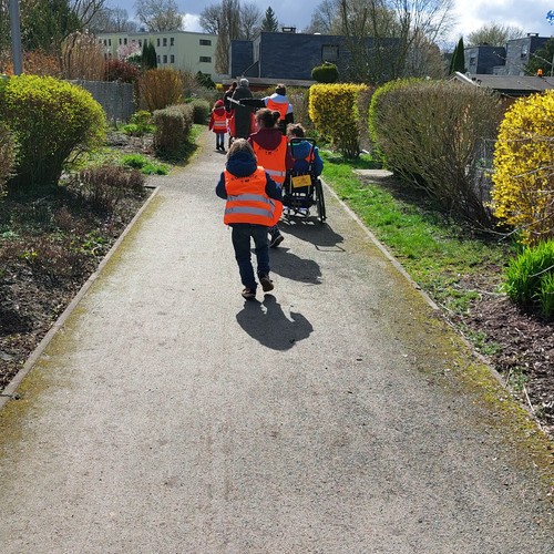 Kinder mit orangenen Warnwesten von hinten zu sehen laufen auf einem Weg durch eine Kleingartenanlage. Eines der Kinder sitzt in einem Rollstuhl.