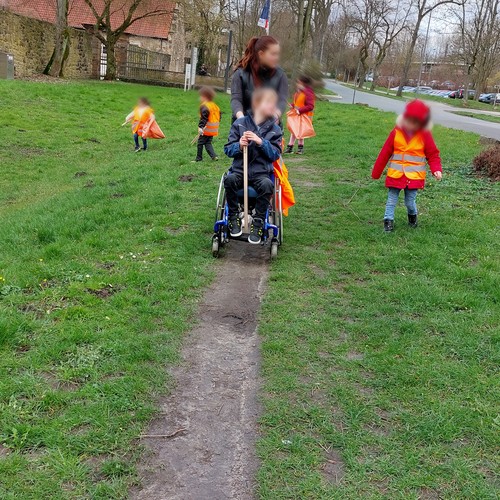 Mehrere Kinder, eines davoin im Rollstuhl, und eine Erwachsene Person mit orangenen Warnwesten sammeln Müll von einer Wiese