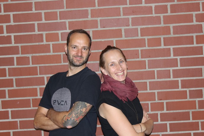 Herr Pluppins und Frau Husmann stehen vor einer gemauerten Wand und lachen.