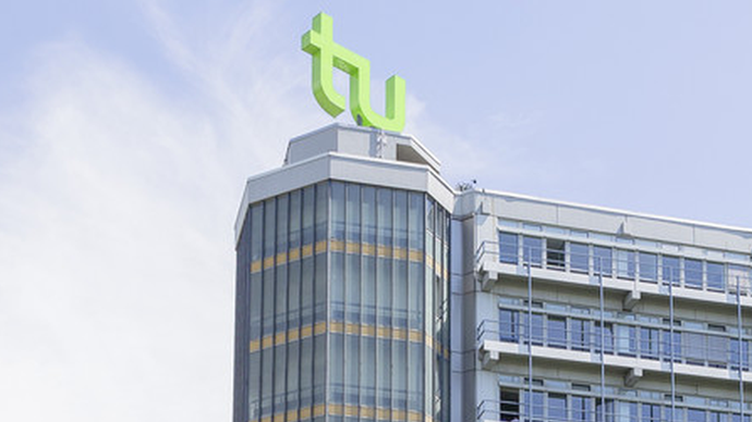 Die oberen Stockwerke eines Gebäudes der TU Dortmund: ein Turm auf gleicher Höhe mit einem sich direkt daran anschließenden Flachdachbau. Auf dem Dach des Turms sind die grünen Buchstaben "TU"