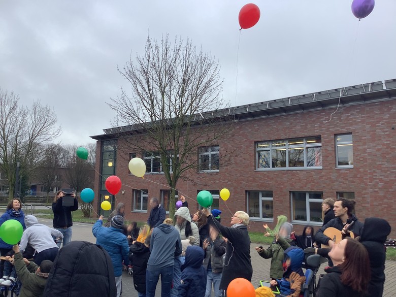 Kinder und Erwachsene stehen vor dem Schulgebäude und lassen bunte Luftballons in den Himnmel steigen