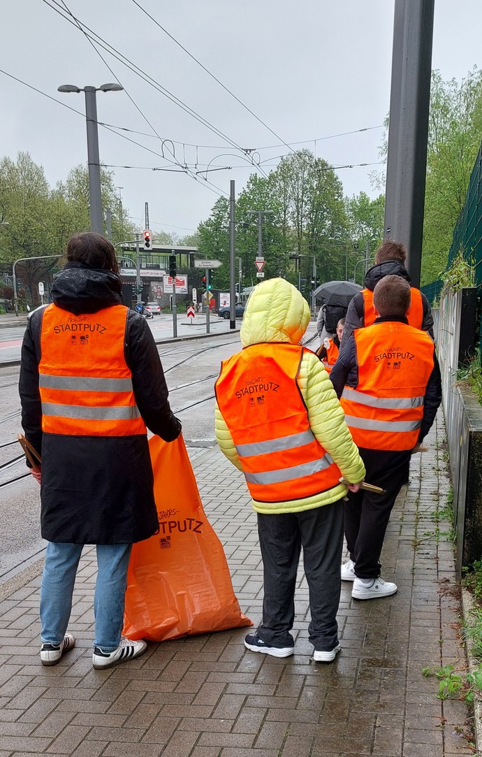 Eine Gruppe Schülerinnen und Schüler mit Warnwesten und Greifzangen stehen an Bahnschienen. Es regnet. Sie sind von hinten zu sehen.
