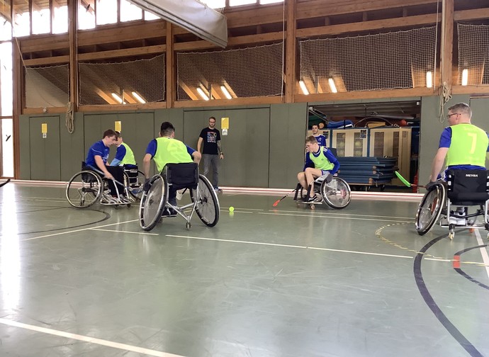 Einige Aktivrollstuhl-Hockeyspielerinnen und Spieler versuchen den Ball zu treffen. Ein Teil der Spieler trägt gelbe Leibchen.