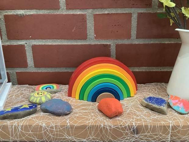 Neben einem Bunten Holzregenbogen liegen mehrere bemalte Steine und daneben steht eine Vase