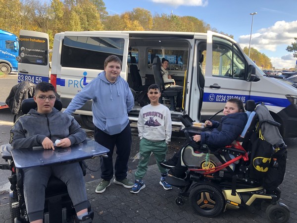 zwei Rollstuhlfahrer und zwei stehende Jungen vor einem Bus in dem bereits ein Rollstuhlfahrer sitzt