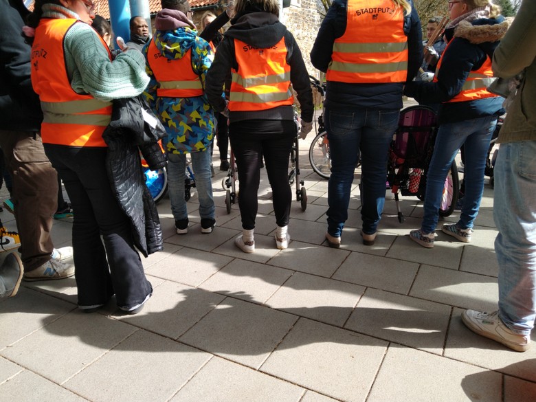 Personen mit orangenen Warnwesten von hinten zu sehen, stehen nebeneinander.