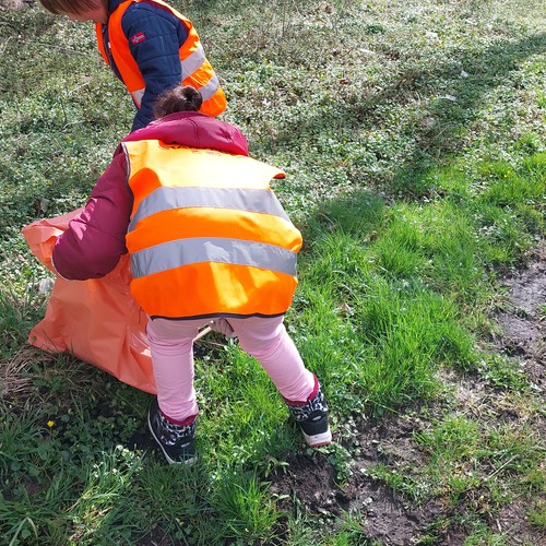 Zwei Kinder mit orangenen Warnwesten von hinten zu sehen bücken sich und sammeln Müll von einer Wiese.