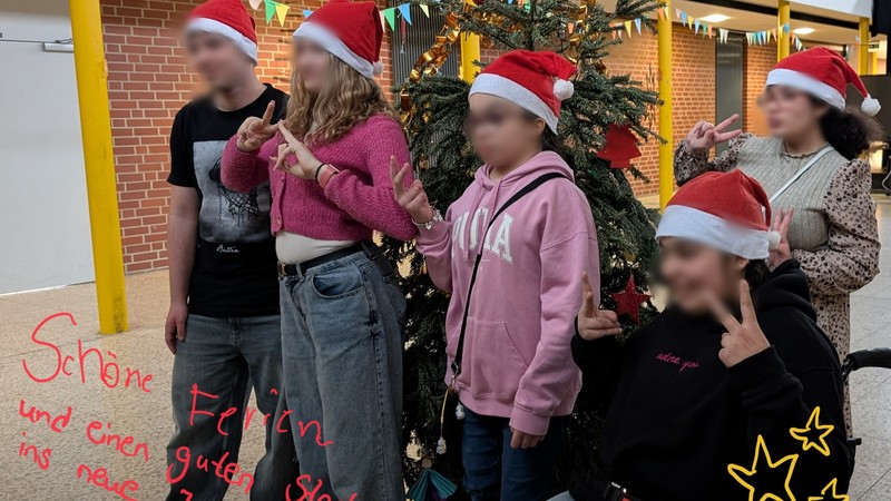 Schülerinnen und Schüler stehen mit Weihnachtsmützen vor dem geschmückten Tannenbaum.
