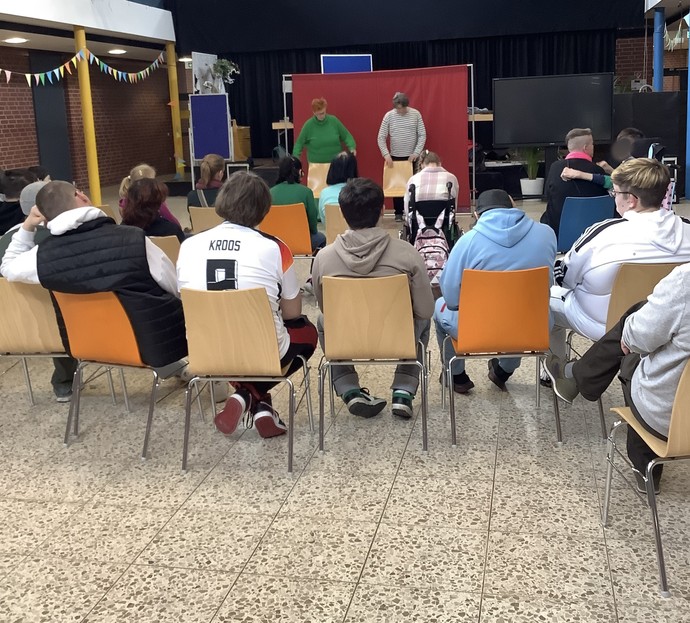 Ein Frau und ein Mann stehen vor einem roten Hintergrund auf der Bühne. Viele Zuschauer/innen schauen zu und sind von hinten abgebildet.