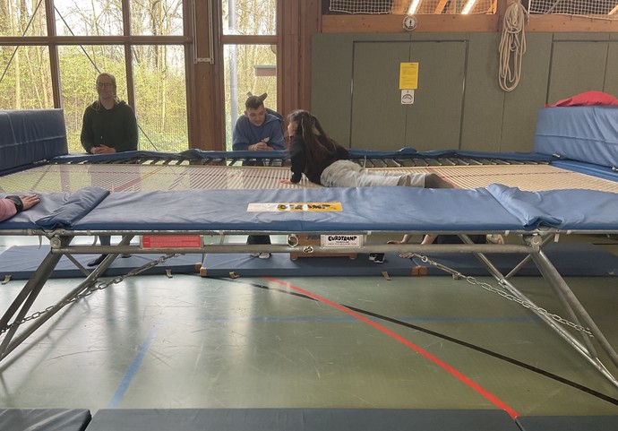 Schülerin liegt auf einem aufgebauten Trampolin. Zwei Erwachsene stehen daneben.