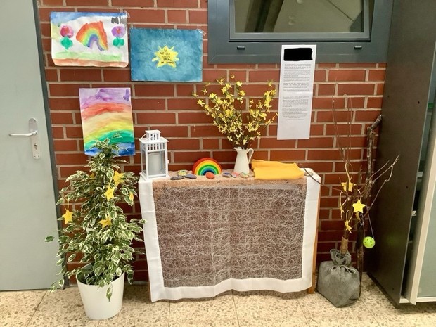 Tisch mit Tischdekce auf dem ein Holzregenbogen steht, daneben bemalte Steine, eine Vase mit gelben Blumen, ein gelbes Tuch und eine Laterne. neben dem Tisch stehen in einer Vase Zweige und eine Topfpflanze, die beide mit Sternanhängern geschmückt sind.
