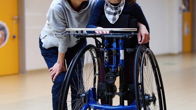 Ein Mädchen im Stehtrainer steht neben einer frau. Beide lächeln sich an.